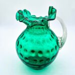 Fenton green hobnail glass pitcher with ruffled rim and clear ribbed handle.
